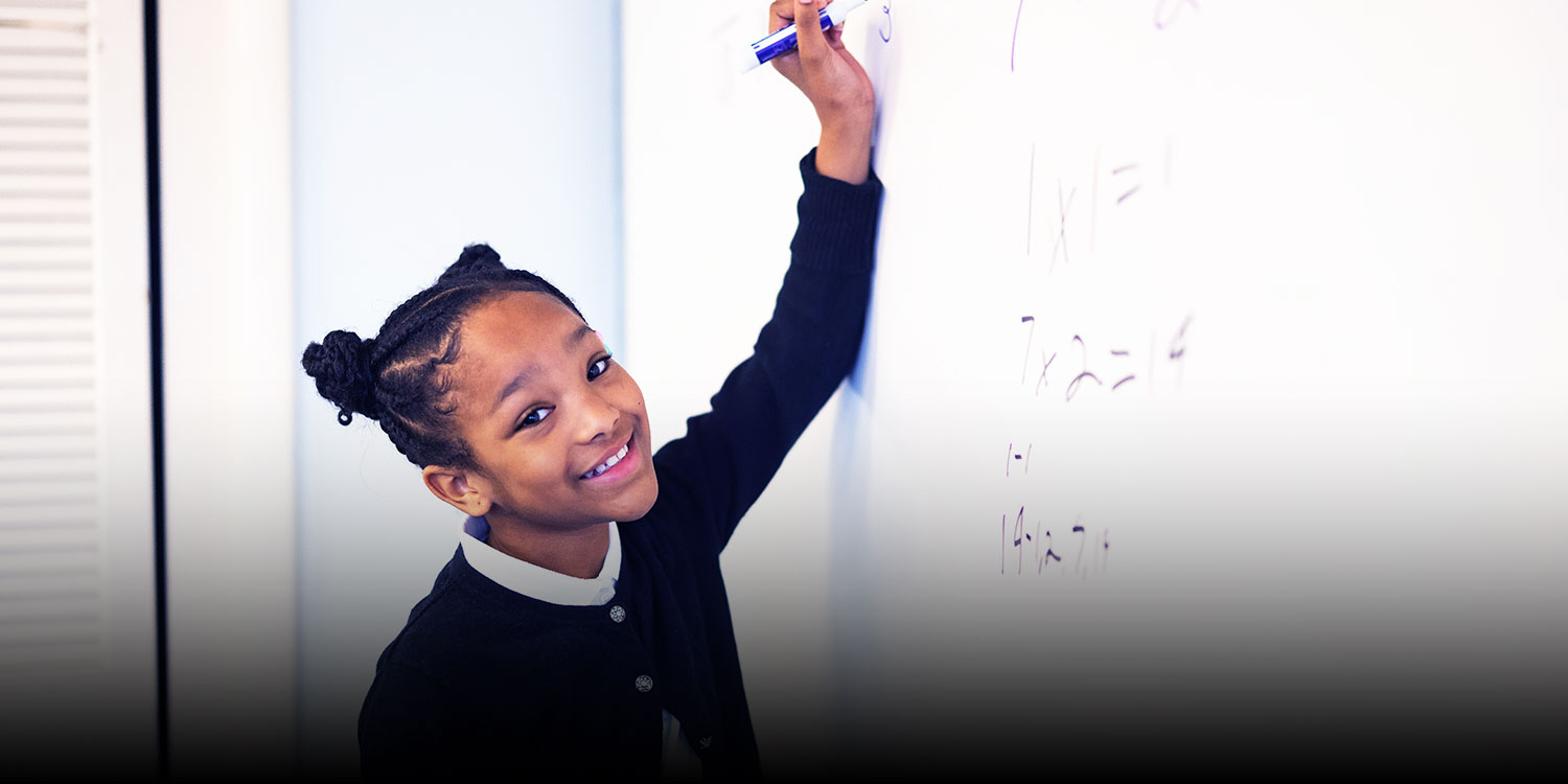 Smiling student writing math on a white board in a classroom.